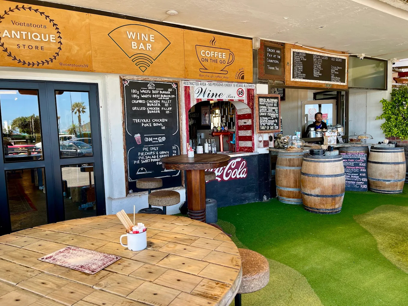 On The Go's beachfront storefront on Beach Road, Gordon's Bay, with chalkboard menus and wine-barrel tables.